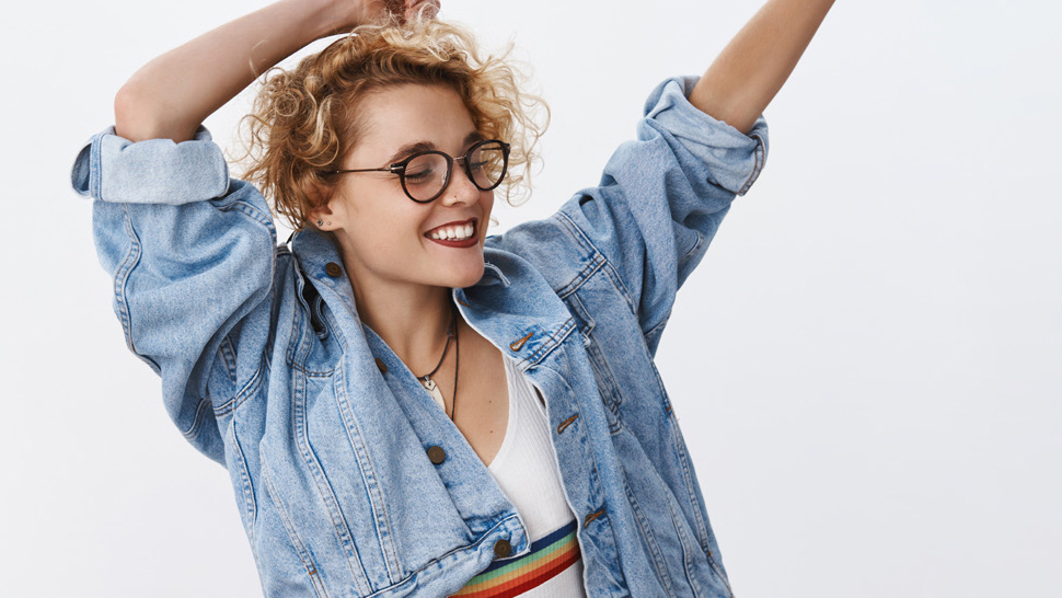 A young girl wearing a denim top and glasses waves her arms and dances happily.